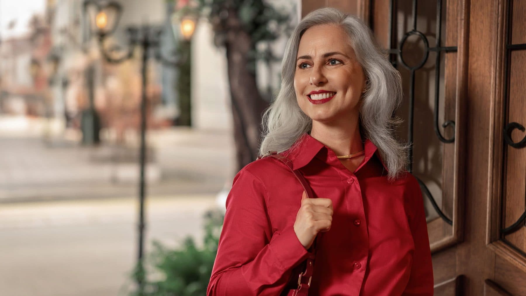 A smiling woman with silver hair wearing a red jacket, standing in a sunny street
